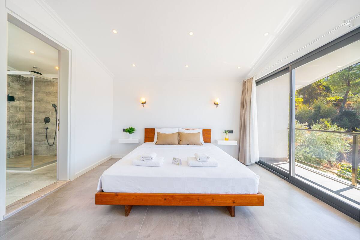 A modern bedroom with a large bed, a glass shower enclosure, and a balcony with a view of trees.