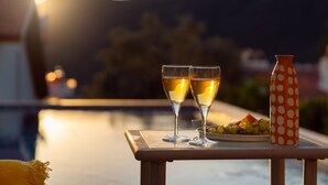 A table with two glasses of a beverage, a plate with grapes and a watermelon, and a polka-dotted bottle, set against a scenic background with a body of water and mountains. - KAYA APARTMENTS KALAMAR (Kas)