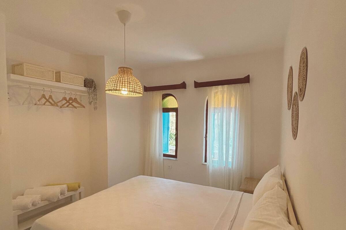 A bedroom with a large bed, a hanging light fixture, a window with curtains, and a wall-mounted shelf with towels.