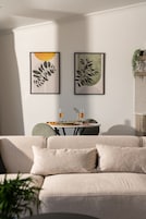A modern living room with a beige sofa, two framed artworks, a small table with drinks, and a hanging plant.
