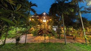 A well-lit building with a central staircase, surrounded by lush greenery and palm trees. - Atoh's Maison (Siem Reap)