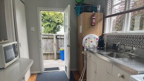 Cocina compacta con microondas, fregadero y estufa. Una puerta da a un espacio exterior con cerca de madera y contenedor azul.