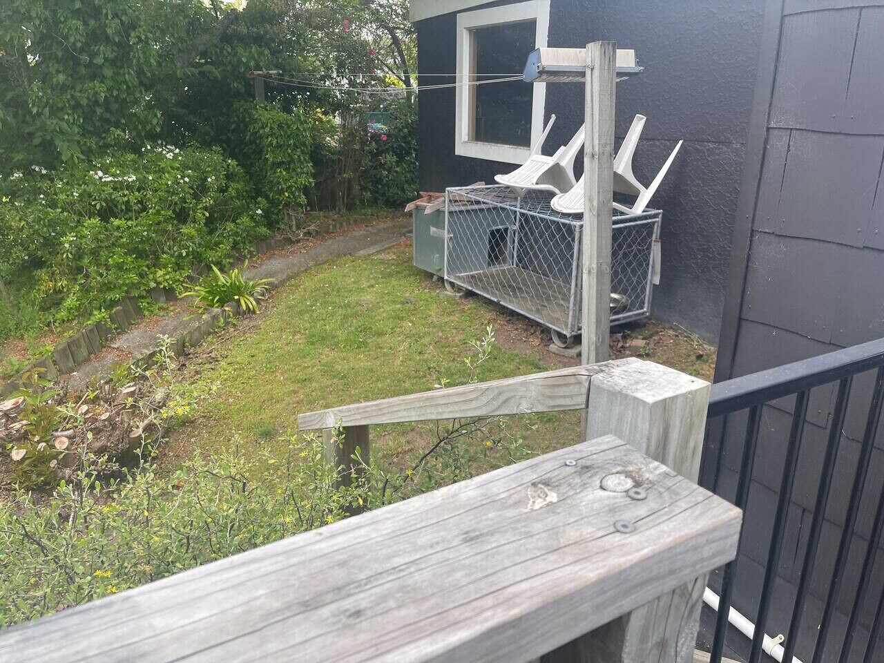 A wooden deck with a metal railing, a small garden with grass and shrubs, and a window with a chair outside.