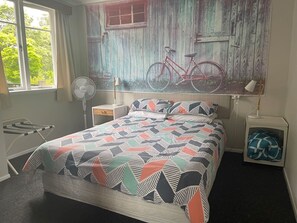 A bedroom with a bed, a fan, a lamp, a small bedside table, and a window with curtains. - Bach 70, Pet Friendly in 2 Mile Bay (Taupō)