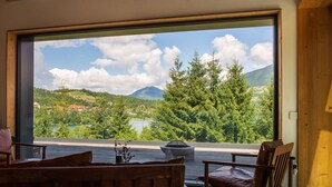A room with a large window offering a view of a lake and mountains. There is a wooden bench and a table with a vase.