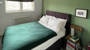 A bedroom with a bed, a radiator, a window with a blind, a bedside table with books, and a wall-mounted picture. - Contemporary 1BD Flat W/balcony - Stoke Newington! (London)