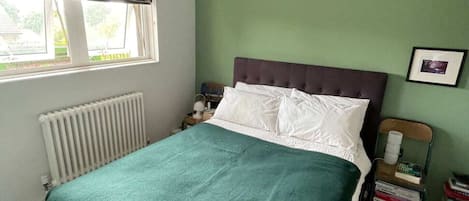 A bedroom with a bed, a radiator, a window with a black blind, a bedside table with a lamp, and a stack of books.