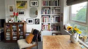 A cozy living room with a wooden table, a bookshelf, and a large window with a view of greenery. - Contemporary 1BD Flat W/balcony - Stoke Newington! (London)