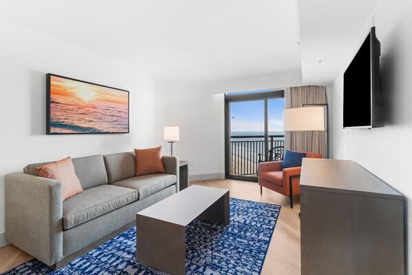 Un salon moderne avec un canapé gris, une table basse et un tapis à motifs bleus. Une grande fenêtre offre une vue sur l’océan, et un balcon est agrémenté d’une chaise.