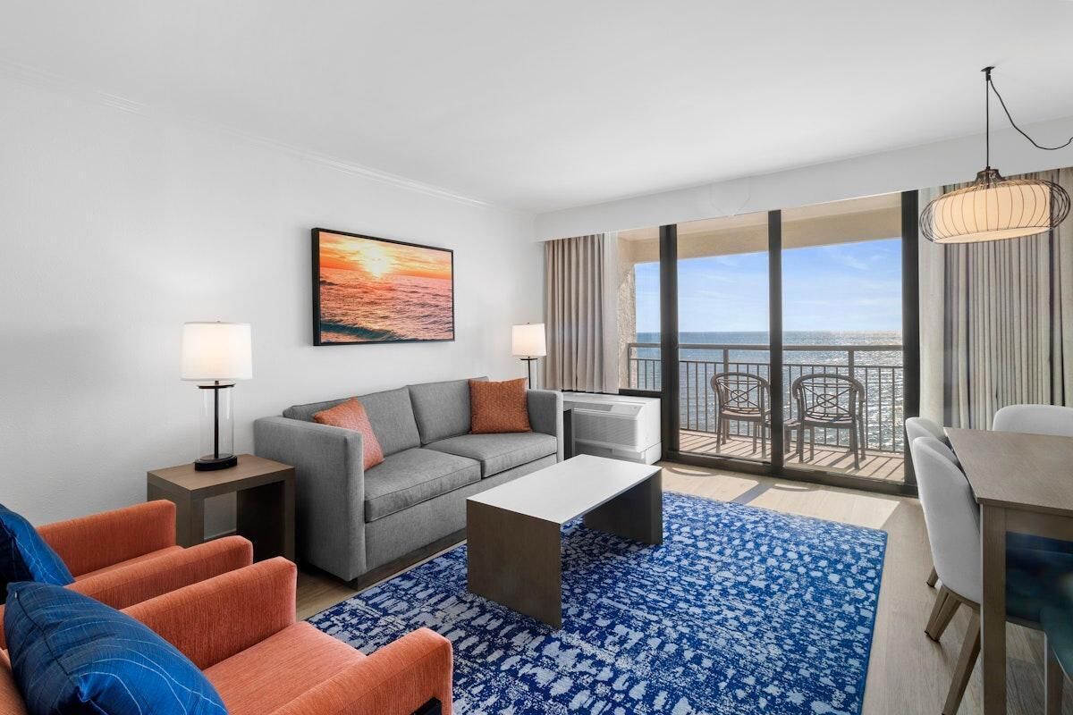 A modern hotel room with a gray sofa, orange armchairs, a coffee table, and a large window offering a view of the ocean.