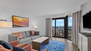 Een moderne hotelkamer met een grijze bank, een blauw kamerkleed, een flatscreen televisie en een balkon met uitzicht op de zee.