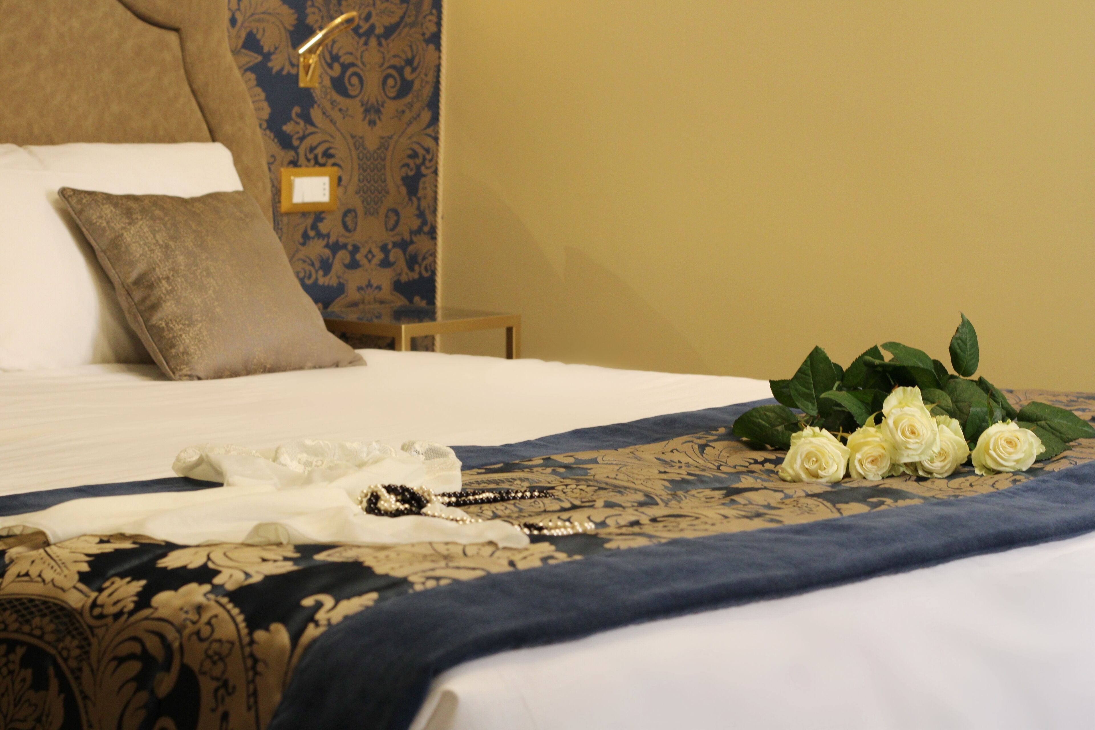 A neatly made bed with a blue and gold patterned bedspread, a white pillow, and a bouquet of white roses.