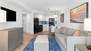Una sala moderna con un sofá gris, una mesa de centro y un televisor. Se observa una cocina al fondo con armarios blancos y una encimera azul.