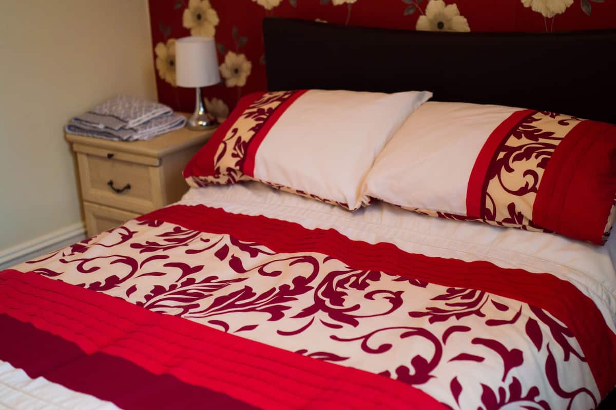 Een keurig opgemaakt bed met een rood-wit gestreept sprei, een passend kussen en een nachtkastje met een lamp en opgevouwen handdoeken.