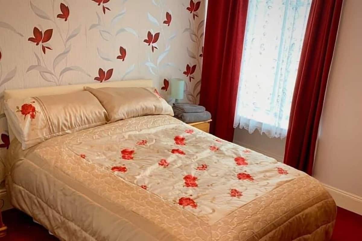 Een net opgemaakt bed met een bloemenbedekking en passende kussens, een nachtkastje met een lamp en een raam met rode gordijnen.