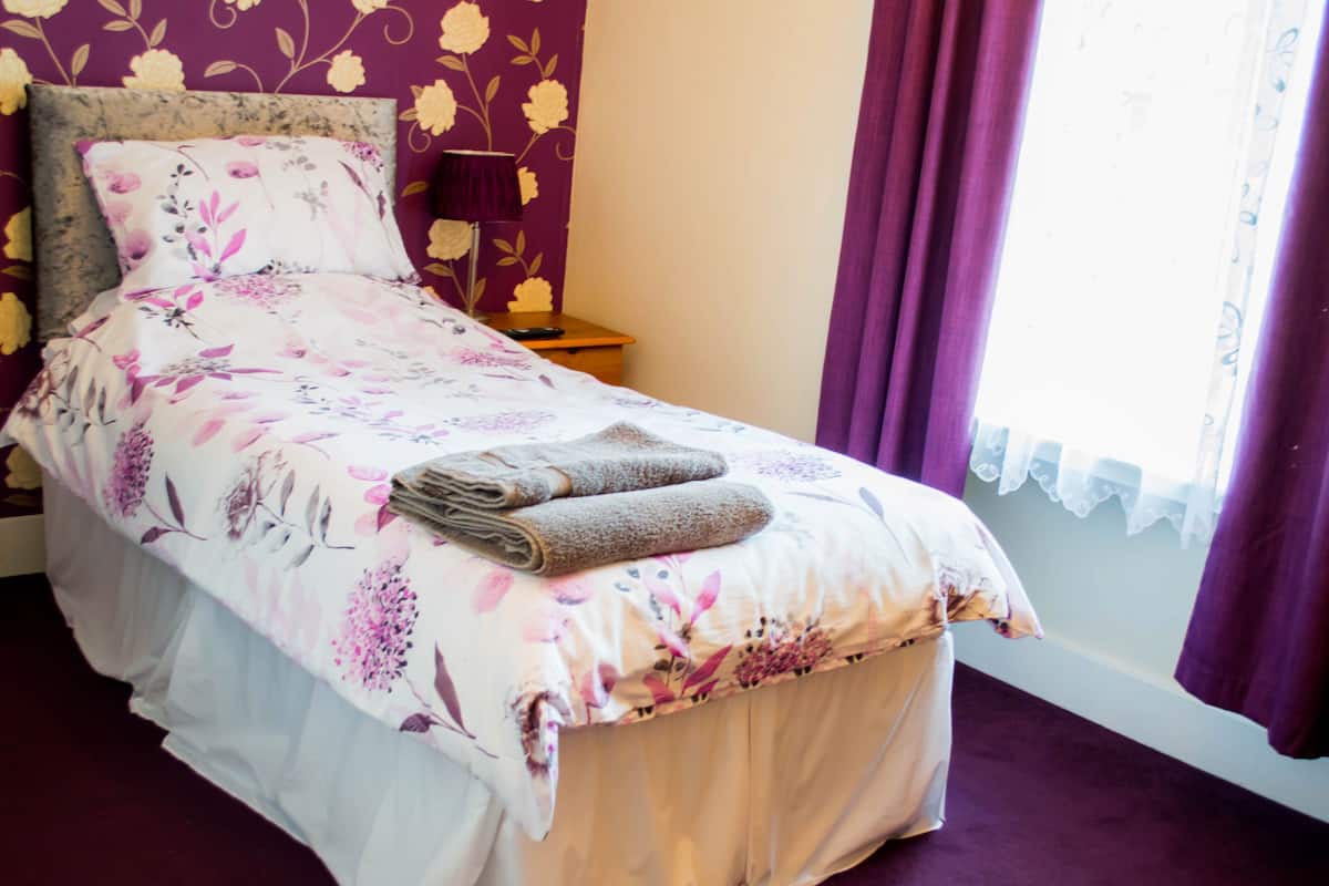 Een net opgemaakt bed met bloemenbedrukking, een nachtkastje met een lamp en een raam met paarse gordijnen.