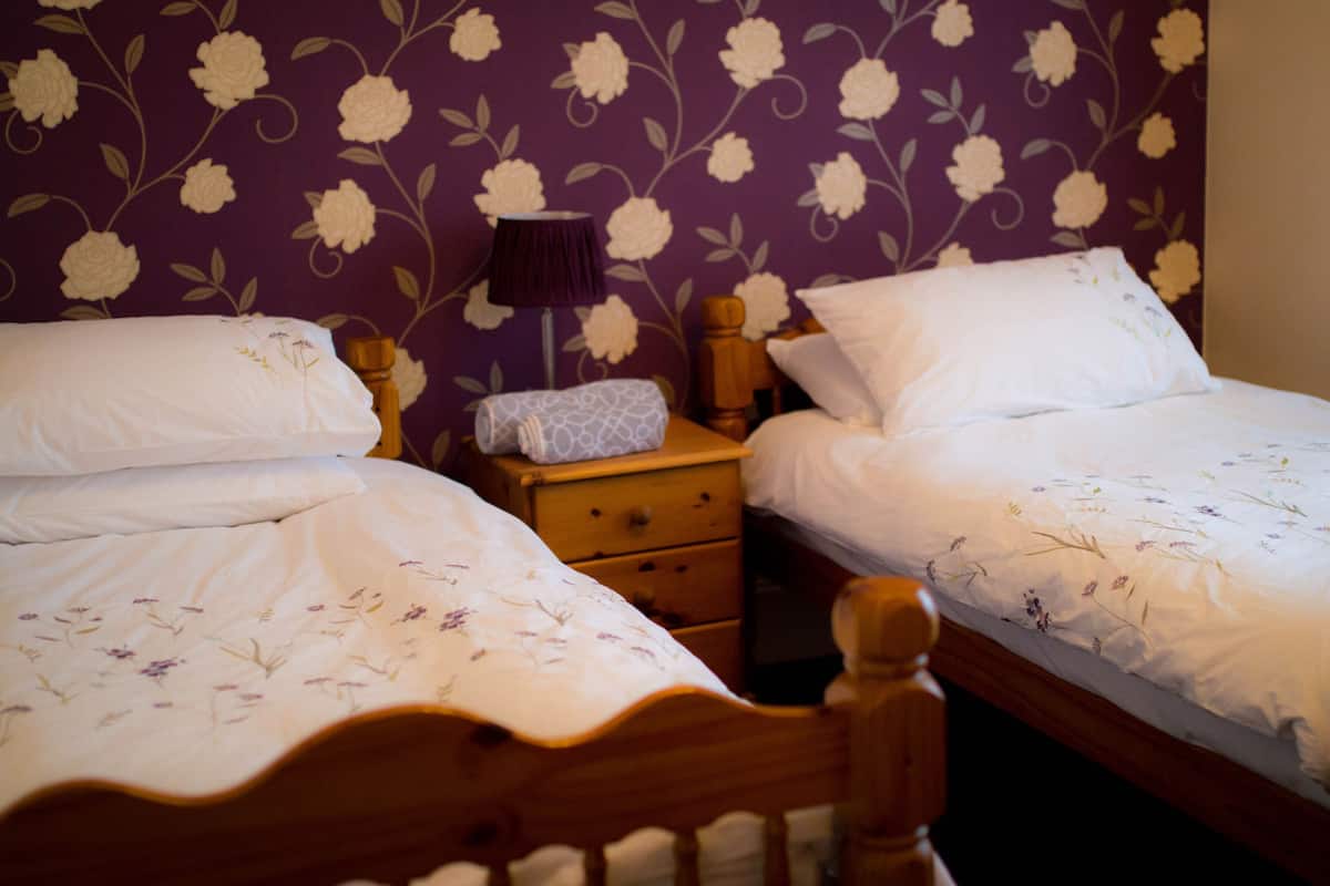 Twee eenpersoonsbedden met houten hoofdeinden, een nachtkastje met een lamp en een wand met bloemenmotief.