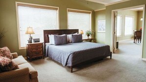 A bedroom with a large bed, a dresser, a nightstand, and a chair. There are two windows with blinds, a ceiling fan, and a door leading to another room.