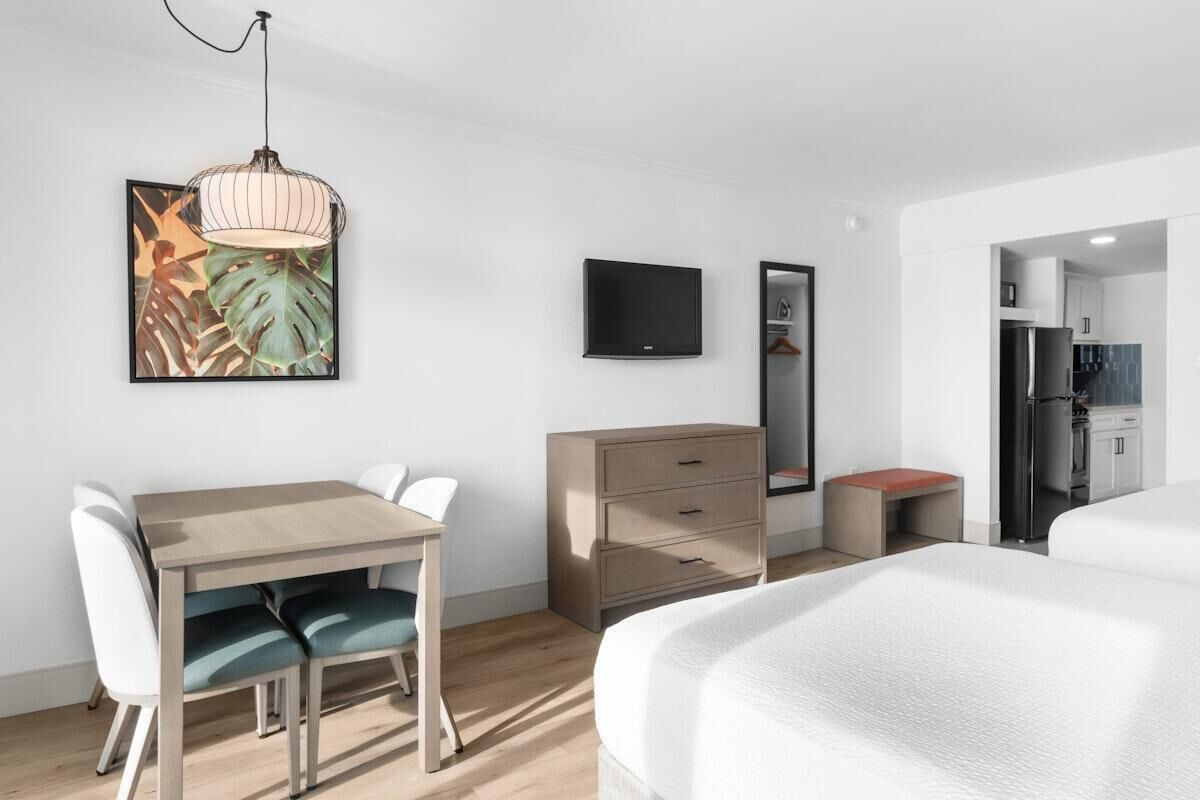 Una habitación de hotel moderna con una cama, un escritorio con sillas, un aparador, un televisor y una cocina visible a través de una puerta abierta.