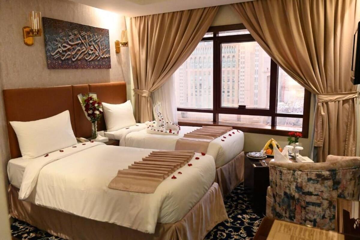 A hotel room with two beds, a large window with curtains, a wall-mounted artwork, and a small table with a vase and flowers.