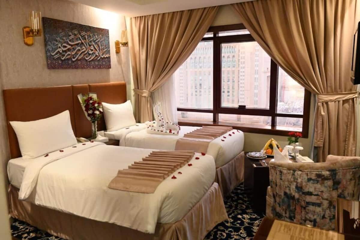 Une chambre d’hôtel avec deux lits, une grande fenêtre avec des rideaux, une œuvre d’art accrochée au mur et une petite table sur laquelle se trouvent un vase et des fleurs.