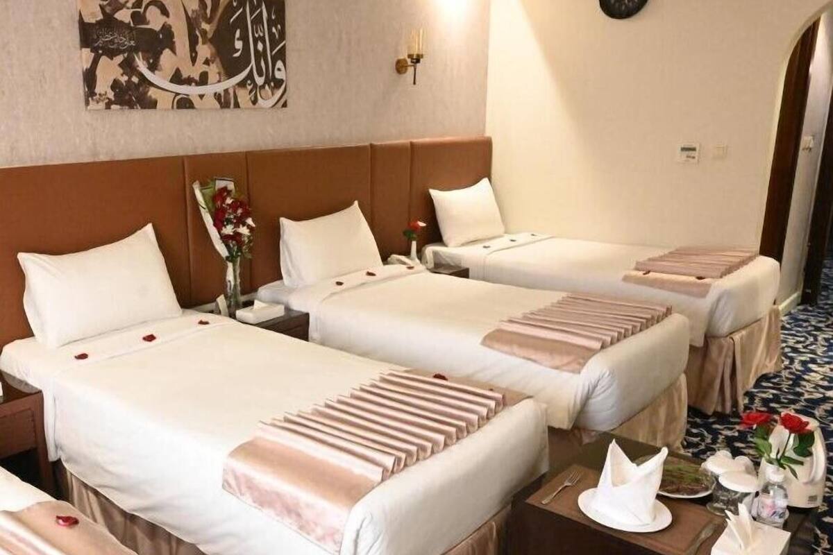 A hotel room with three beds, each with a white bedspread and a pink decorative pillow. There is a clock on the wall and a vase with flowers on the nightstand.