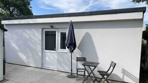 Un petit bâtiment blanc avec une porte et des fenêtres, une terrasse avec une table et des chaises, et un parapluie sombre.