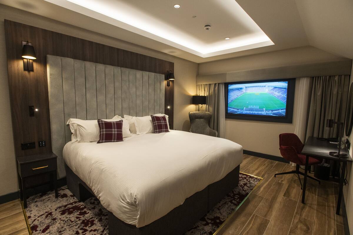 A modern hotel room with a large bed, a desk, a TV, and a view of a stadium.