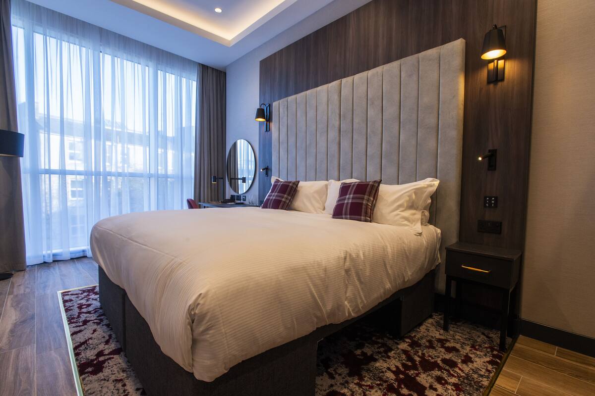 A modern hotel room with a large bed, a bedside table, a nightstand, a mirror, and a view of the outdoors through sheer curtains.