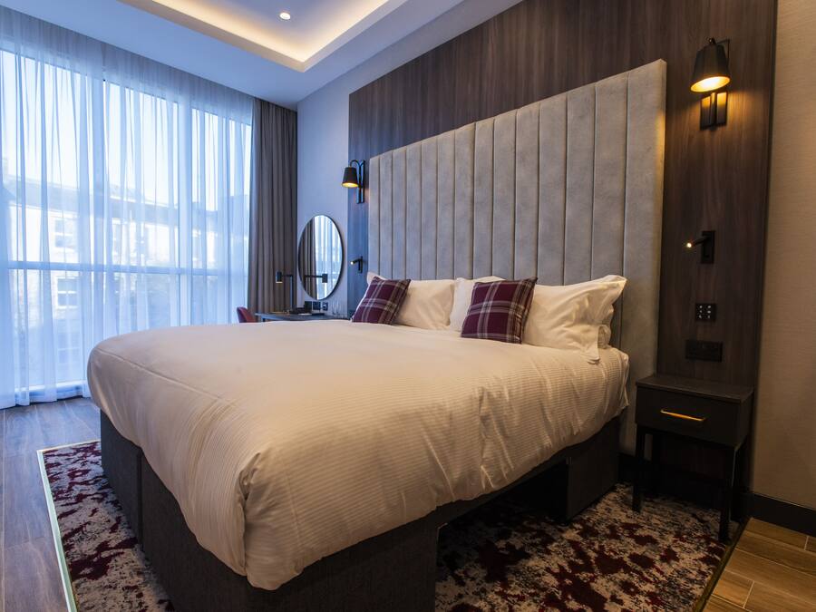 A modern hotel room with a large bed, a bedside table, a nightstand, a mirror, and a view of the outdoors through sheer curtains.