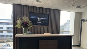 En receptionsdisk med en tv-skærm der viser et bybillede, en vase med blomster og et skilt der angiver et business center.