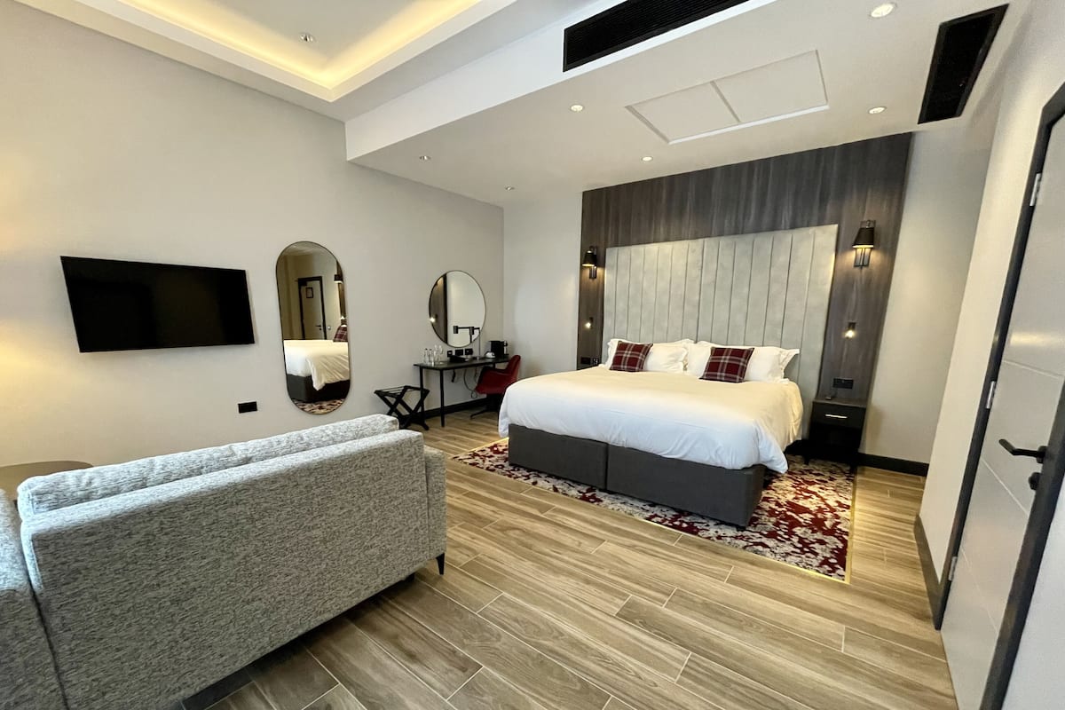 A modern bedroom with a large bed, a grey sofa, a flat-screen TV, a desk, and a mirror.