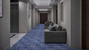 A hallway with a blue patterned carpet, a sofa with pillows, framed pictures on the wall, and a door with a number sign. - Shaza Regency Plaza (Madinah)