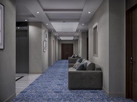 A hallway with a blue patterned carpet, a sofa with pillows, framed pictures on the wall, and a door with a number sign.