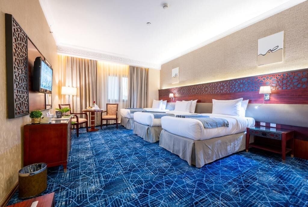 A hotel room with a blue patterned carpet, two beds, a desk with a chair, a TV, and a decorative headboard.