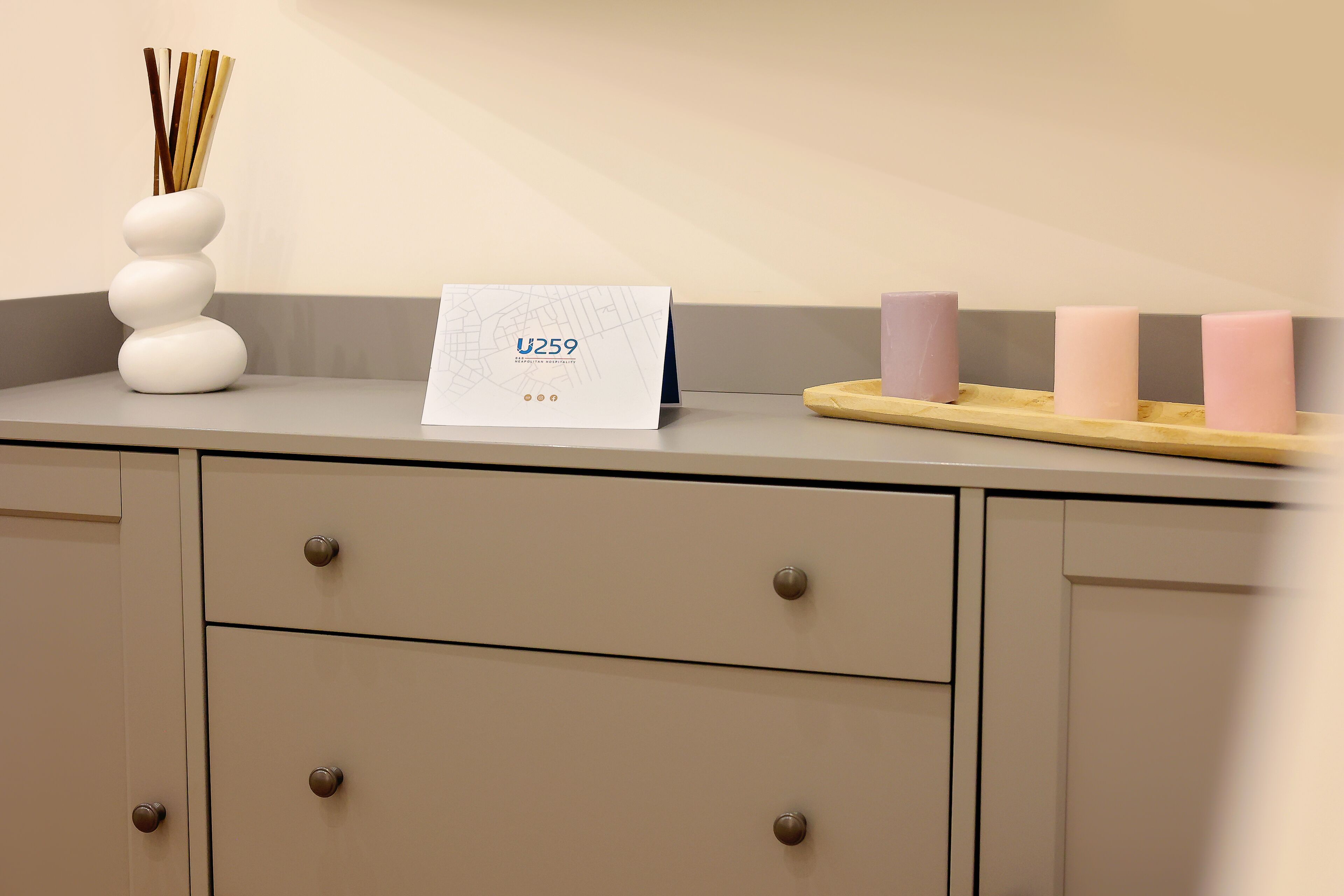 A reception desk with a sign reading "U259," a white vase with sticks, and three pink candles on a wooden tray.