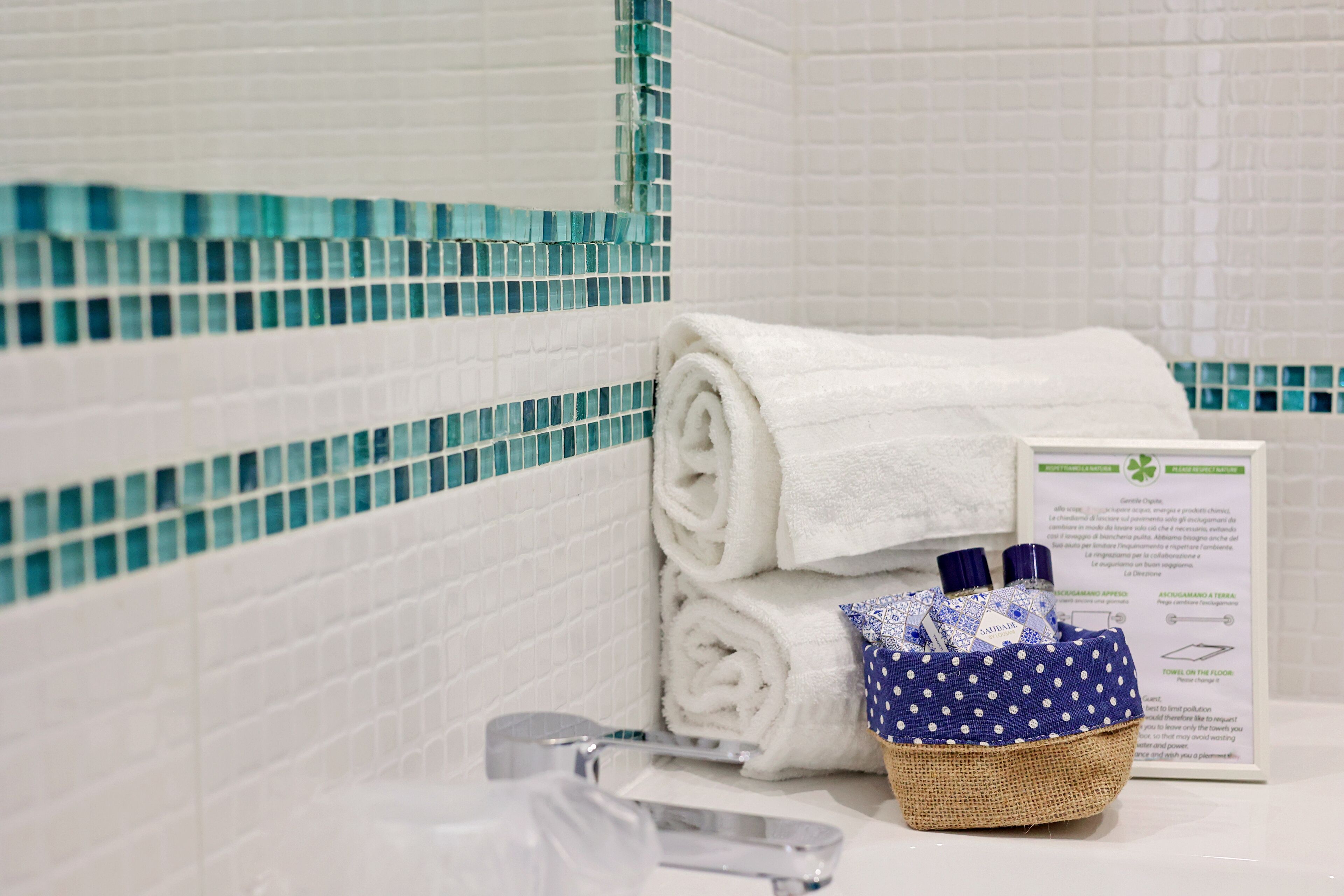 A bathroom with white tiled walls, a blue mosaic border, a rolled white towel, a wicker basket with toiletries, and a faucet.