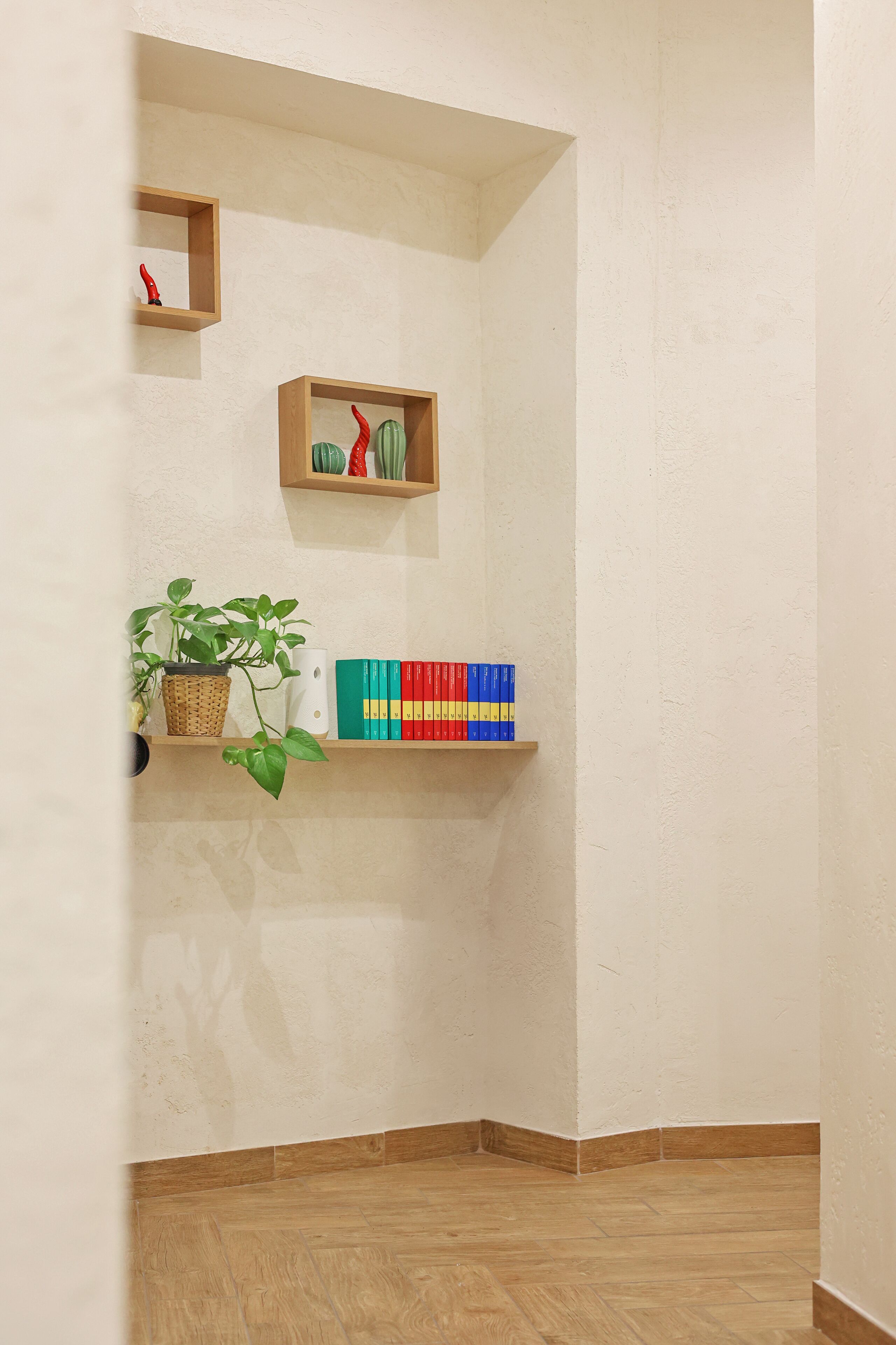 A corner of a room with a wooden floor, a white textured wall, and shelves with a plant, books, and decorative items.