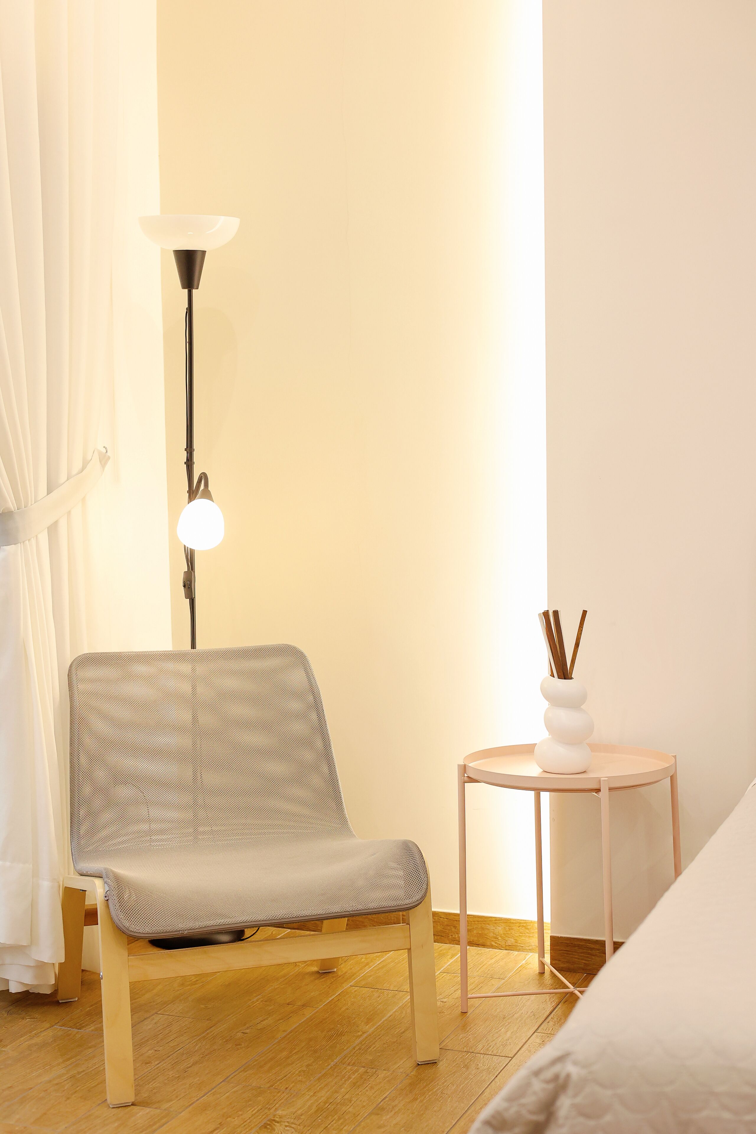 Une pièce avec un plancher en bois, un fauteuil recouvert d’un tissu gris, une petite table ronde surmontée d’un vase blanc et un lampadaire.