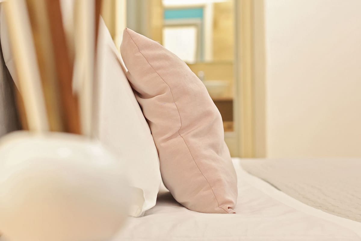 A neatly made bed with a white and beige pillow, a wooden headboard, and a view of a room with a wooden door and a blue chair.