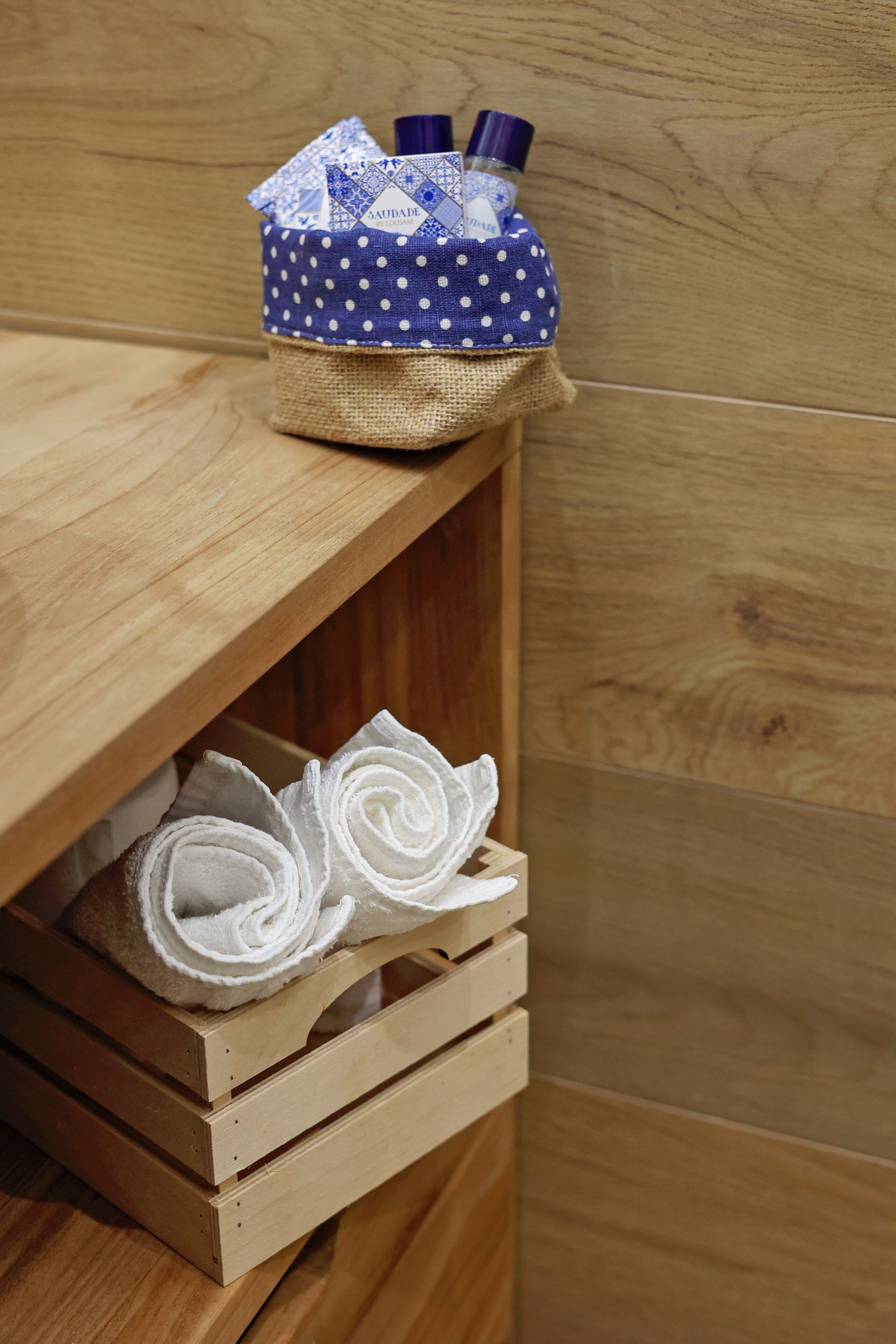 A wooden shelf with a blue polka-dotted basket containing two bottles and a rolled white towel.
