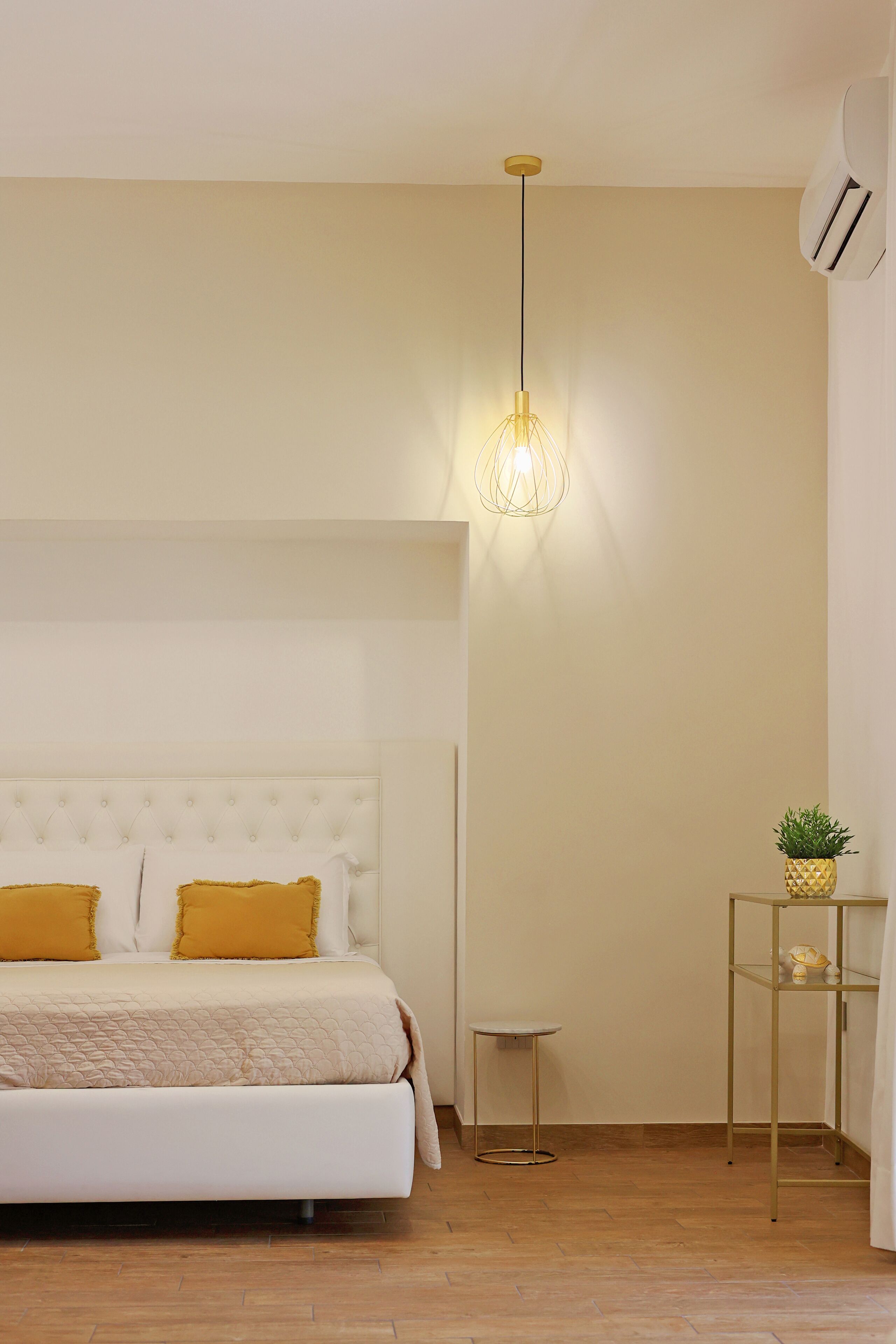 A bedroom with a bed, a hanging light fixture, a small table, and a shelf with a plant.