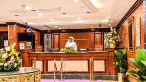 Un hall d'hôtel avec un comptoir d'accueil en bois, un homme en uniforme blanc et un grand arrangement floral.