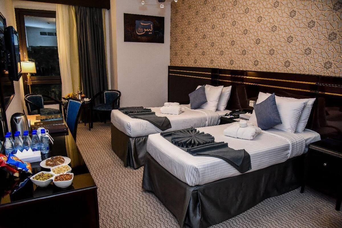A hotel room with two beds, a desk with a lamp, chairs, and a view of the outside.