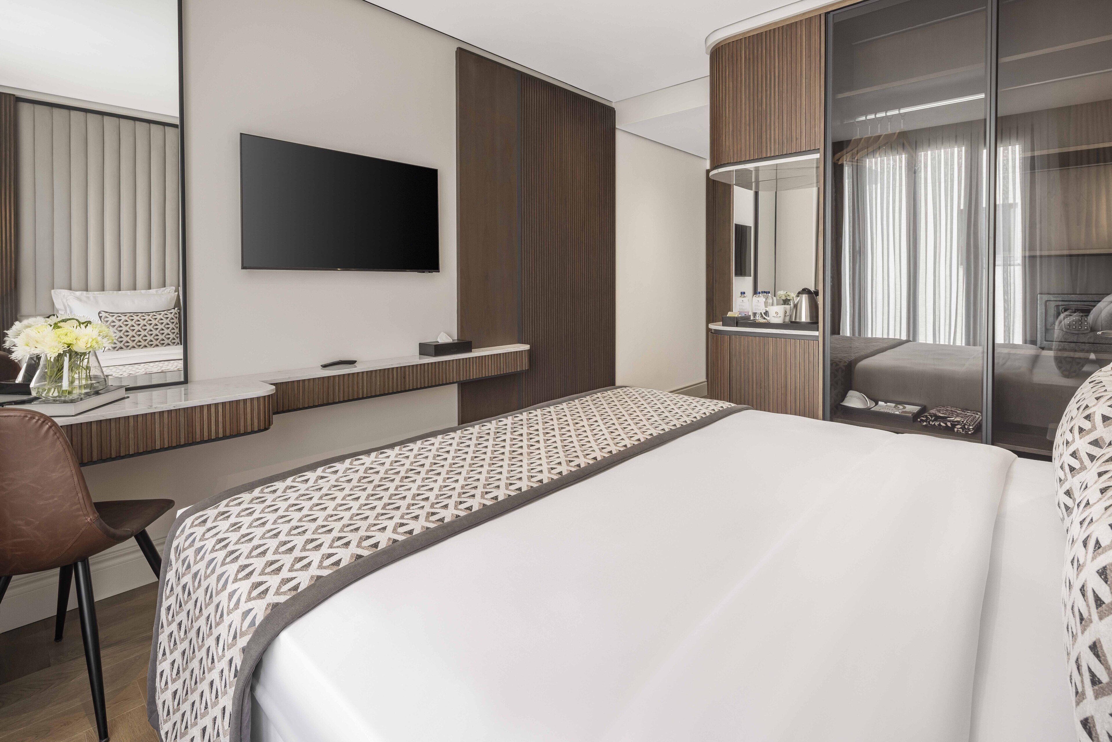 Une chambre d’hôtel moderne dotée d’un grand lit, d’un téléviseur à écran plat fixé au mur, d’un bureau avec une chaise et d’une porte-fenêtre coulissante donnant sur un balcon.