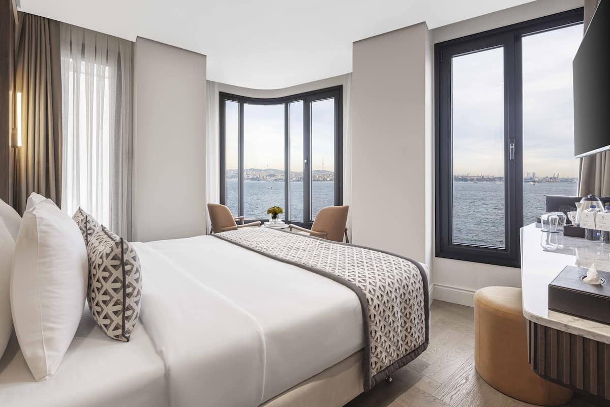 Ein modernes Hotelzimmer mit einem großen Bett, zwei Sesseln, einem kleinen Tisch mit einer Vase und Blick auf das Wasser sowie die Stadtlandschaft durch große Fenster.