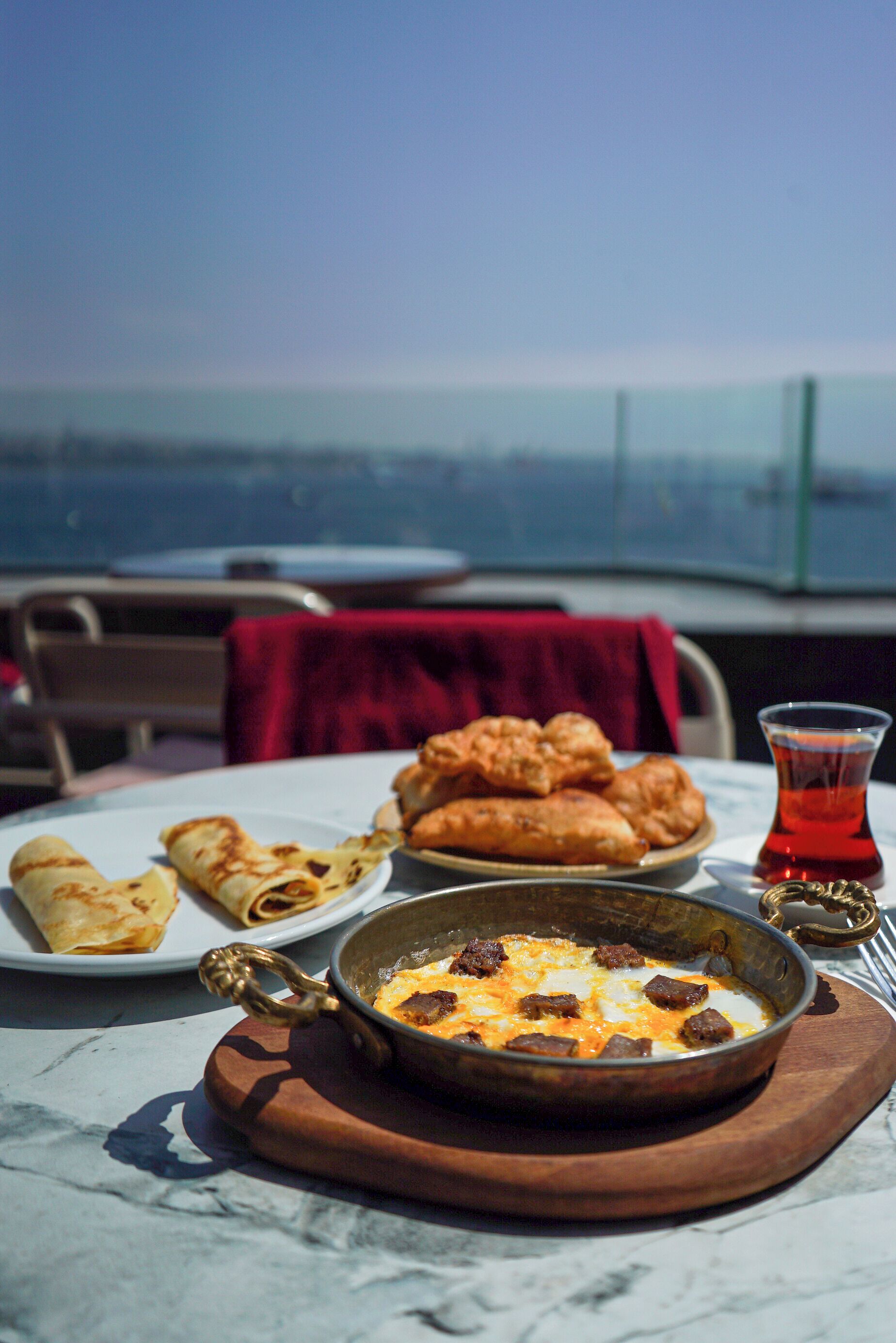 Une table dressée avec un repas composé d’un bol de soupe, d’une assiette de pâtisseries et d’un verre de thé, le tout mis en valeur par un ciel dégagé et une vue lointaine en arrière-plan.