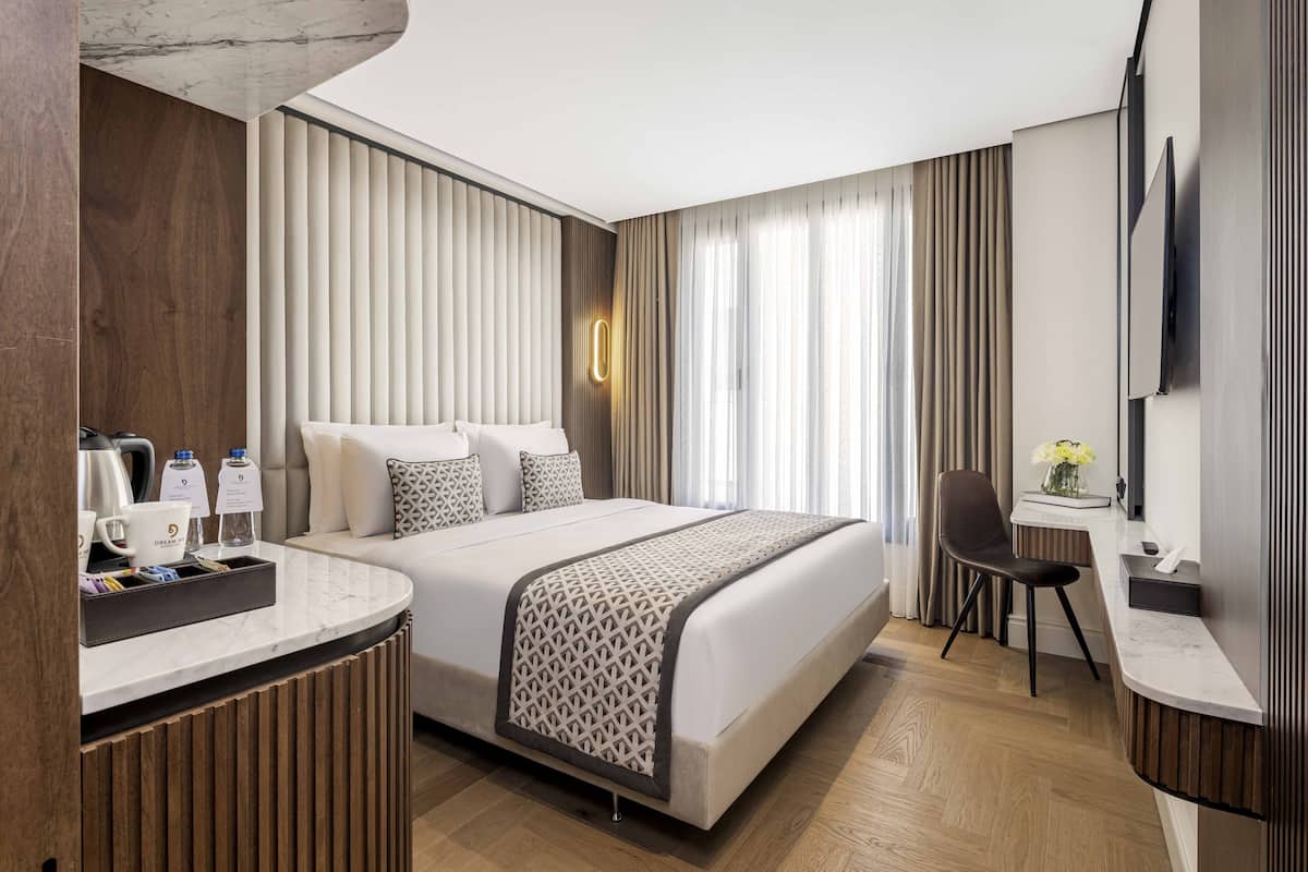 Ein modernes Hotelzimmer mit einem großen Bett, einem Schreibtisch und einem Stuhl. Auf dem Schreibtisch befinden sich eine Kaffeemaschine, eine Wasserflasche und eine Vase mit Blumen.