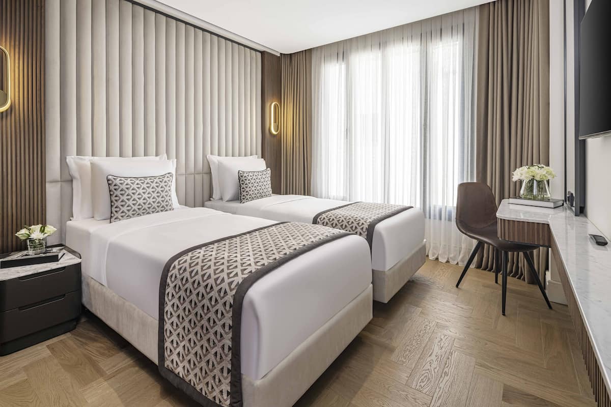 Ein Hotelzimmer mit zwei Betten, einem Schreibtisch, einem Stuhl, einem Fernseher und einem Nachttisch mit Blumenvase.