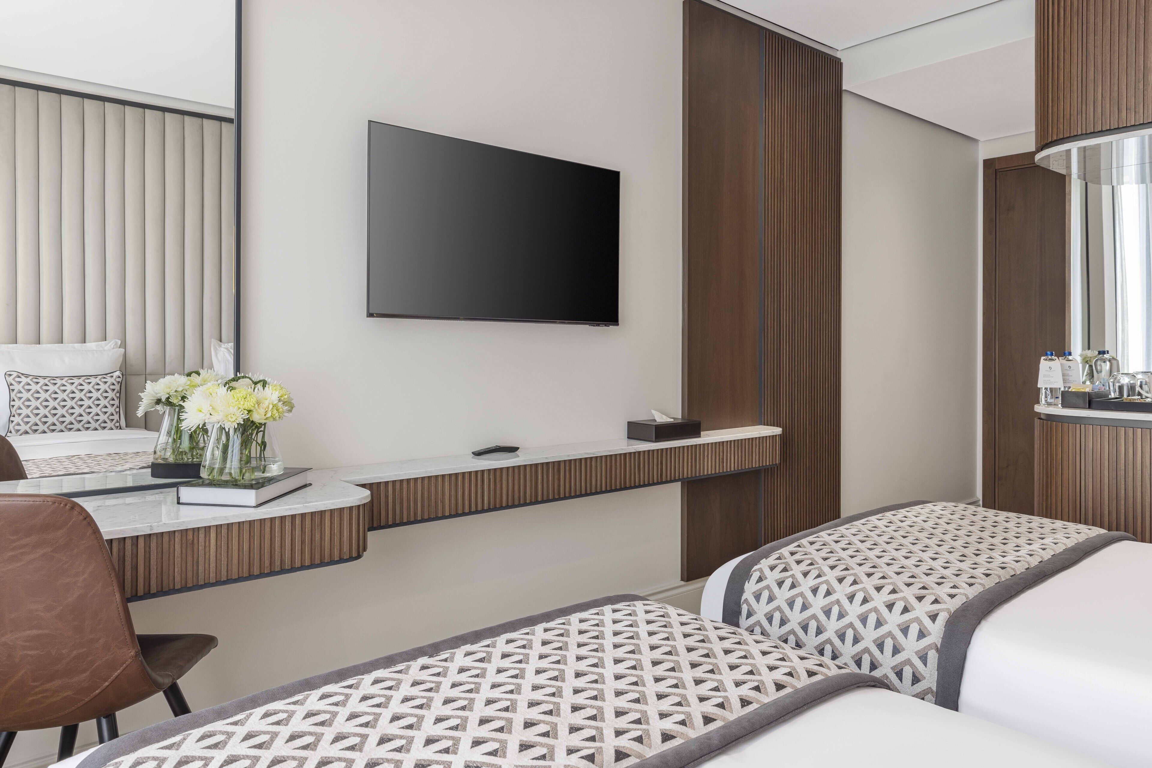 Une chambre d’hôtel moderne dotée d’un téléviseur à écran plat fixé au mur, d’un petit bureau agrémenté d’un vase de fleurs et de deux lits avec une literie à motifs.